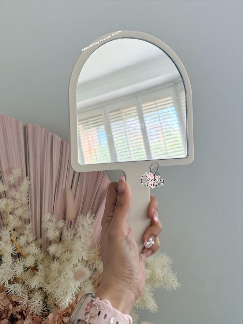 Puede incluir: Un espejo de mano color crema con la parte superior arqueada se sostiene frente a una pared gris claro. El mango del espejo est&aacute; integrado en su marco. La direcci&oacute;n del sitio web "www.ceejaycreations.com.au" est&aacute; impresa en la parte superior del espejo.