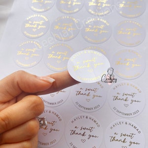 以下が含まれることがあります： 「a sweet thank you」とハートと日付「22 September 2023」が書かれた金色の文字の白い丸いステッカー。ステッカーは結婚式用です。