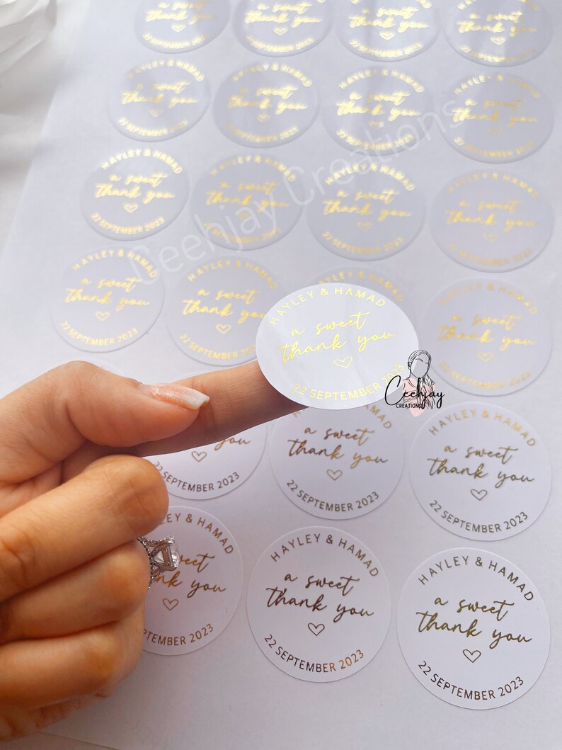 以下が含まれることがあります： 「a sweet thank you」とハートと日付「22 September 2023」が書かれた金色の文字の白い丸いステッカー。ステッカーは結婚式用です。