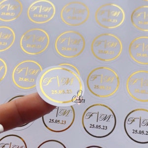 以下が含まれることがあります： 金色の文字が書かれた白い丸いステッカーのシート。ステッカーには、「F」と「M」の文字が縦線で区切られており、「25.05.23」の日付が書かれています。