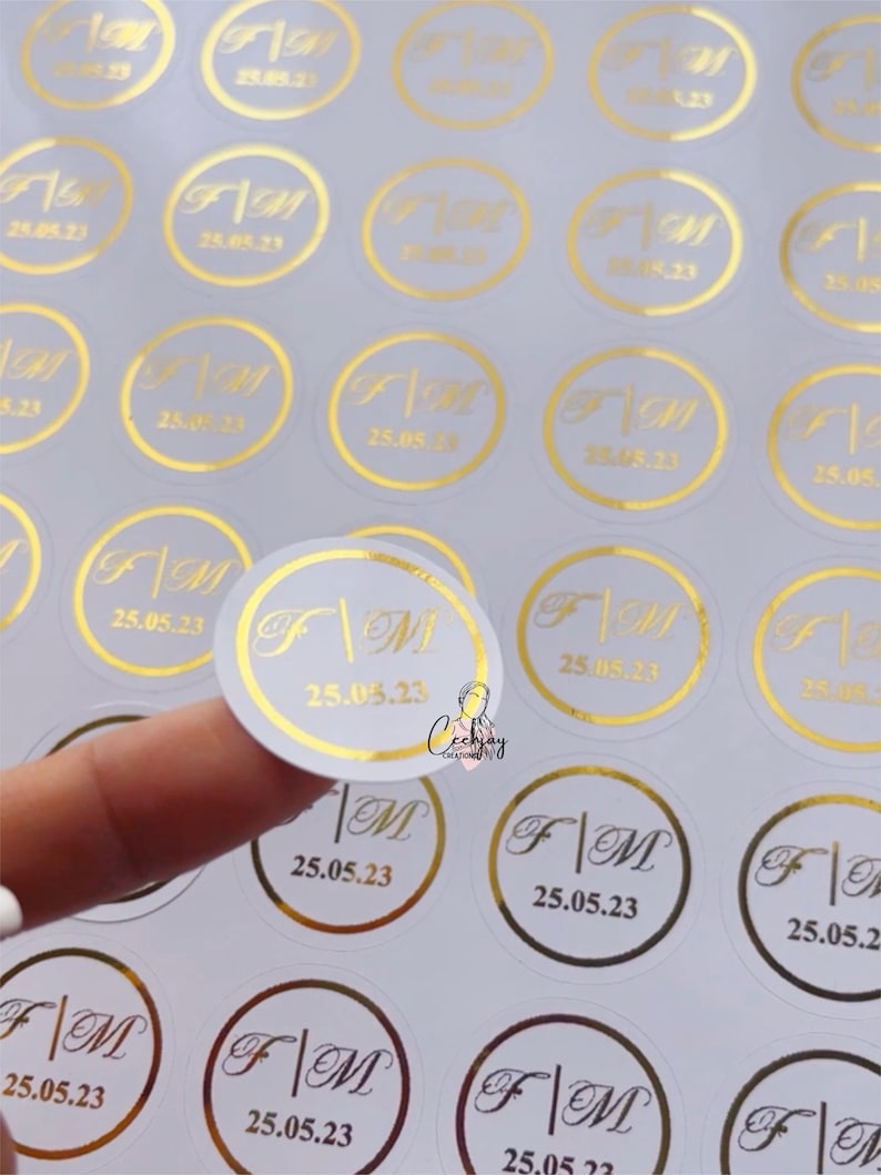 以下が含まれることがあります： 金色の文字が書かれた白い丸いステッカーのシート。ステッカーには、「F」と「M」の文字が縦線で区切られており、「25.05.23」の日付が書かれています。