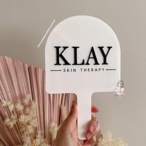 Puede incluir: Un letrero blanco con asa, con el texto negro "KLAY SKIN THERAPY". El letrero tiene una parte superior redondeada y se sostiene frente a un arreglo floral rosa y beige. La direcci&oacute;n del sitio web www.ceehjaycreations.com.au est&aacute; impresa en el letrero.