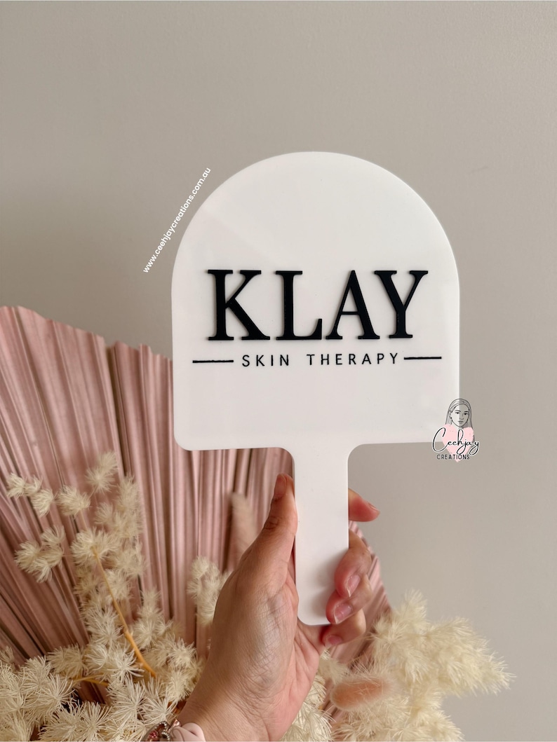 Puede incluir: Un letrero blanco con asa, con el texto negro "KLAY SKIN THERAPY". El letrero tiene una parte superior redondeada y se sostiene frente a un arreglo floral rosa y beige. La direcci&oacute;n del sitio web www.ceehjaycreations.com.au est&aacute; impresa en el letrero.