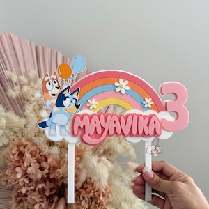 Pu&ograve; includere: Un topper per torta rosa e blu con un arcobaleno, due cani dei cartoni animati e il nome "Mayavika" con il numero "3" sul lato.