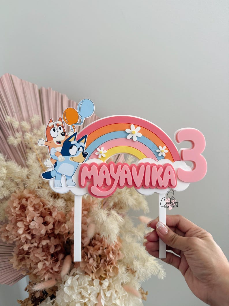 Pu&ograve; includere: Un topper per torta rosa e blu con un arcobaleno, due cani dei cartoni animati e il nome "Mayavika" con il numero "3" sul lato.