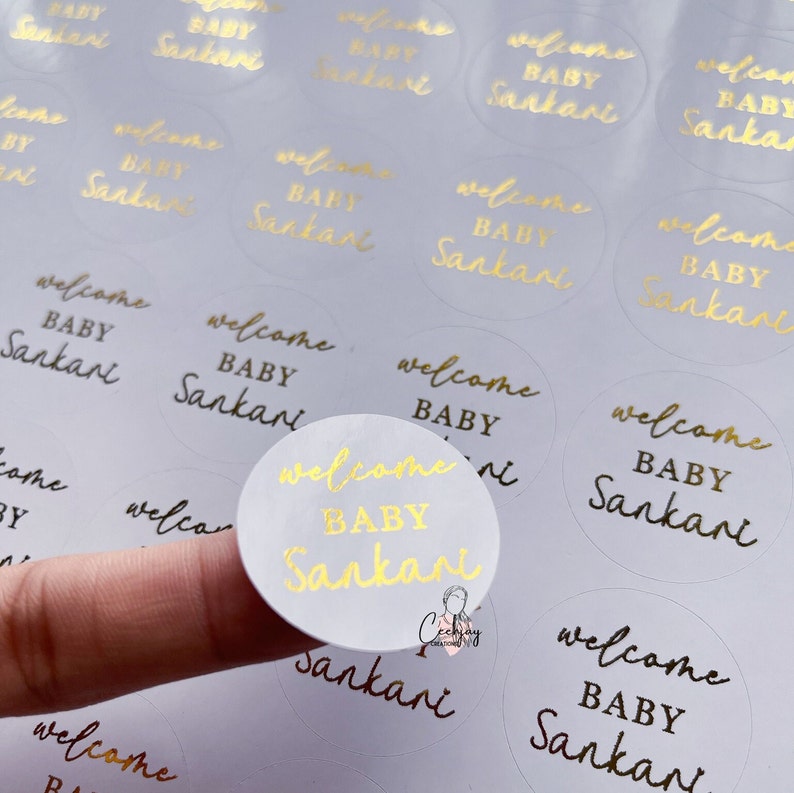 以下が含まれることがあります： 白い円形のラベルに「Welcome Baby Sankari」という金箔の文字が書かれたゴールドフォイルステッカー。