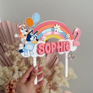 Pu&ograve; includere: Un topper per torta arcobaleno rosa e blu con due cani dei cartoni animati, una nuvola con il nome "Sophie" e il numero "2".