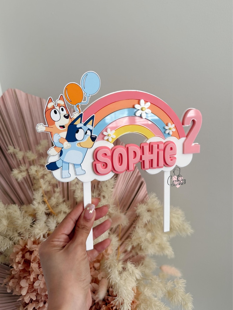 Pu&ograve; includere: Un topper per torta arcobaleno rosa e blu con due cani dei cartoni animati, una nuvola con il nome "Sophie" e il numero "2".