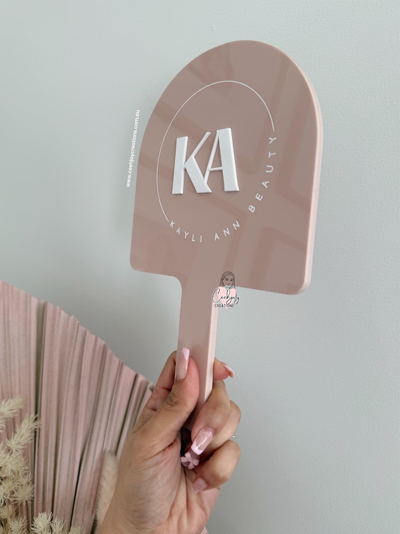 Puede incluir: Un letrero rosa p&aacute;lido con un asa, que muestra el texto blanco "KA" dentro de un c&iacute;rculo, con las palabras "KAYLI ANN BEAUTY" que lo rodean. El letrero lo sostiene una persona. El letrero tambi&eacute;n tiene el texto "ceehjoycreations.com.au".