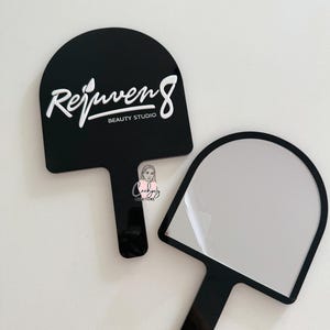 Puede incluir: Dos espejos de mano negros. Uno tiene el texto blanco "Rejuven8 BEAUTY STUDIO". El otro espejo tiene una superficie reflectante. Ambos tienen una parte superior redondeada y un mango.