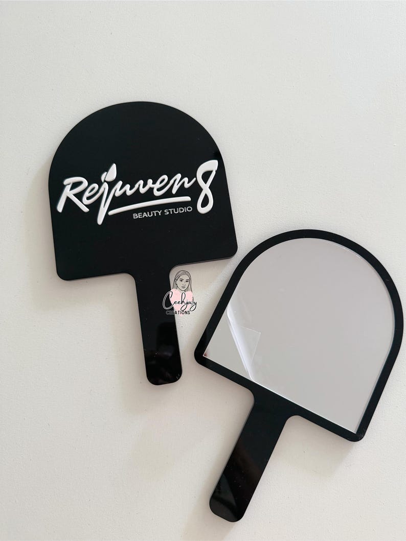 Puede incluir: Dos espejos de mano negros. Uno tiene el texto blanco "Rejuven8 BEAUTY STUDIO". El otro espejo tiene una superficie reflectante. Ambos tienen una parte superior redondeada y un mango.