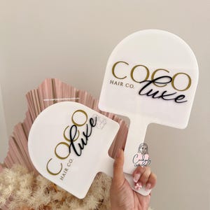 Puede incluir: Dos letreros blancos en forma de paleta con el texto "COCO LUXE HAIR CO." en dorado y negro. Una persona sostiene un letrero. Los letreros est&aacute;n frente a un arreglo floral rosa y beige.