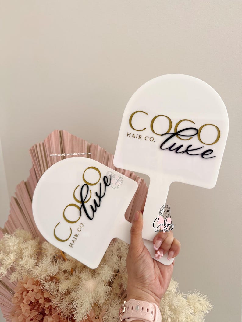Puede incluir: Dos letreros blancos en forma de paleta con el texto "COCO LUXE HAIR CO." en dorado y negro. Una persona sostiene un letrero. Los letreros est&aacute;n frente a un arreglo floral rosa y beige.
