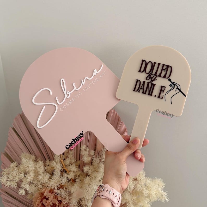Puede incluir: Dos paletas rosas para arte de tatuaje cosm&eacute;tico. Una paleta tiene el nombre "Sibina" en escritura blanca y el texto "Cosmetic Tattoo Art". La otra paleta tiene el texto "Dolled by Dani.E" y una ilustraci&oacute;n de un pincel de maquillaje. Ambas tienen un mango.