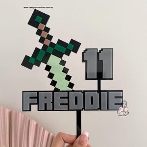 May include: A Minecraft-style cake topper with a pixelated sword, the name "FREDDIE" in grey block letters, and the number "11". The topper is black with green, brown, and light green details. The website address "www.ceehjaycreations.com.au" is visible.