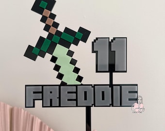 Puede incluir: Un adorno para tarta con temática de Minecraft que presenta una espada pixelada, el nombre "FREDDIE" en letras grises y el número "11". El adorno es negro con detalles verdes, marrones y verde claro. La dirección del sitio web "www.ceehjaycreations.com.au" es visible.