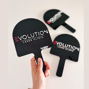 Puede incluir: Carteles negros en forma de paleta con el texto "EVOLUTION LASER CLINIC" en blanco y rojo. Una mano sostiene un cartel. Los carteles tienen un asa y una parte superior redondeada. Los carteles est&aacute;n sobre una superficie blanca.