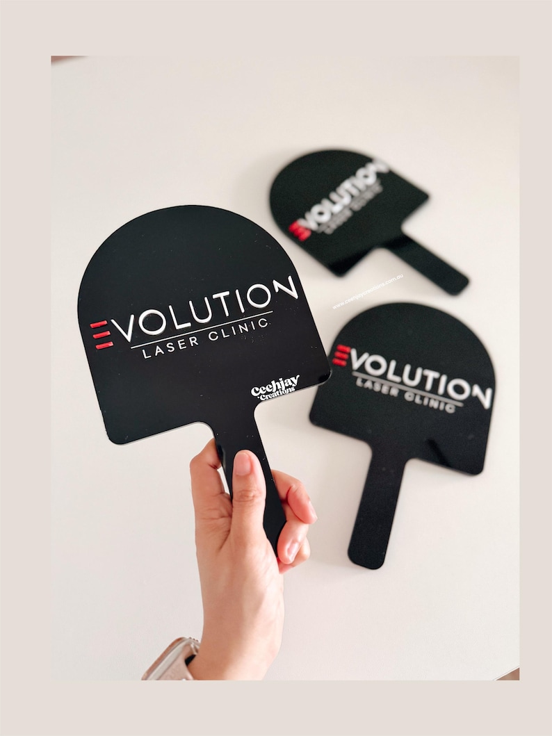 Puede incluir: Carteles negros en forma de paleta con el texto "EVOLUTION LASER CLINIC" en blanco y rojo. Una mano sostiene un cartel. Los carteles tienen un asa y una parte superior redondeada. Los carteles est&aacute;n sobre una superficie blanca.