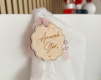 Puede incluir: Una etiqueta de calcetín navideño de madera con el nombre "Amarah Star" grabado en una elegante escritura. La etiqueta tiene un borde festoneado y está sujeta a un calcetín blanco y esponjoso con una cinta rosa. El fondo incluye una figura de Papá Noel.