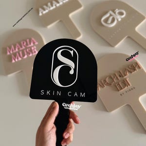 Puede incluir: Una mano sostiene un letrero negro con el texto "SKIN CAM" y un logotipo "S" blanco. Otros letreros beige en forma de paleta muestran nombres como "AMARIE" y "MARIA MUSE".