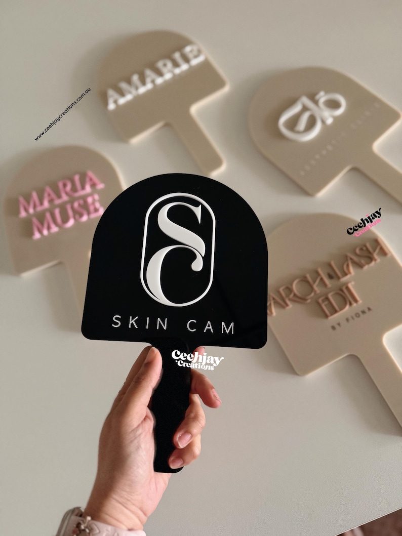 Puede incluir: Una mano sostiene un letrero negro con el texto "SKIN CAM" y un logotipo "S" blanco. Otros letreros beige en forma de paleta muestran nombres como "AMARIE" y "MARIA MUSE".