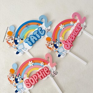 Pu&ograve; includere: Tre topper per torte di Bluey con archi di arcobaleno, nuvole e i nomi Tate, Spencer e Sophie. Ogni topper presenta un personaggio di Bluey che tiene un palloncino.