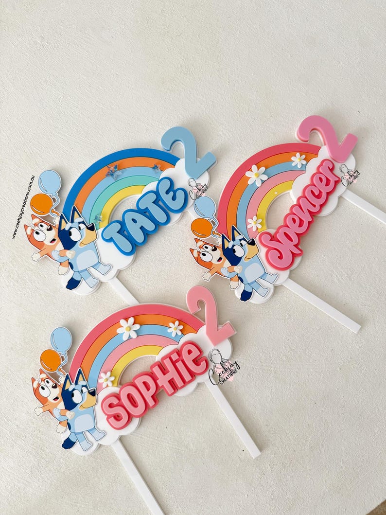 Pu&ograve; includere: Tre topper per torte di Bluey con archi di arcobaleno, nuvole e i nomi Tate, Spencer e Sophie. Ogni topper presenta un personaggio di Bluey che tiene un palloncino.