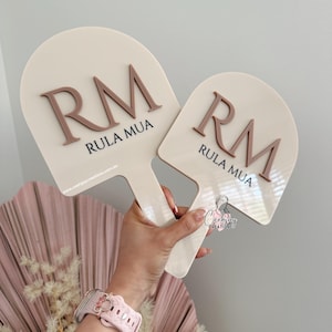 May include: Two off-white paddle-shaped signs with brown lettering. The signs have the letters "RM" and the words "RULA MUA" printed on them. The signs are held in a person's hand. The background includes a pink fan-shaped decoration and dried flowers.