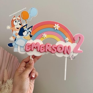 May include: A pink and blue cake topper with a rainbow, two cartoon dogs, and the name "EMERSON" and the number "2".