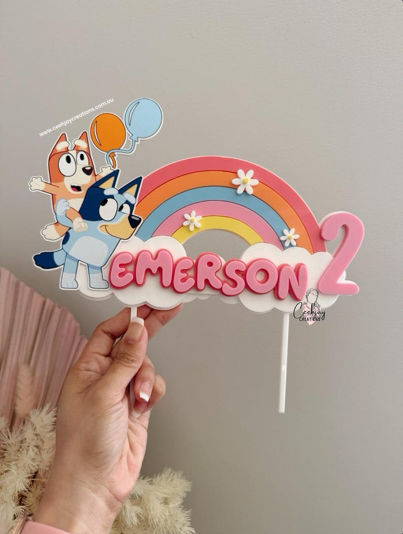 Pu&ograve; includere: Un topper per torta rosa e blu con un arcobaleno, due cani dei cartoni animati e il nome "EMERSON" e il numero "2".
