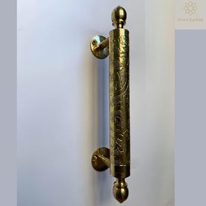 May include: A brass door pull with an ornate, engraved design. The pull is cylindrical with decorative end caps and mounting hardware. The surface is polished and reflects light, showcasing the intricate patterns. The text "Brass Kashbah" is in the upper right corner.