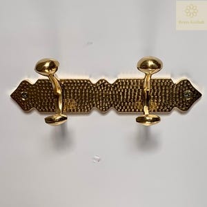 Puede incluir: Un perchero de pared dorado con dos ganchos. El perchero tiene un diseño de metal martillado texturizado y está montado con tornillos. Los ganchos son redondeados y tienen un diseño decorativo y ornamentado. El perchero es de latón.