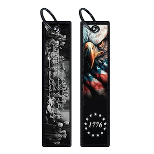 May include: Two black keychains with a white background. The keychain on the left features a black and white image of the signing of the Declaration of Independence. The keychain on the right features a colorful image of an American bald eagle with the American flag draped over it. The text "1776" is printed in white below the eagle.