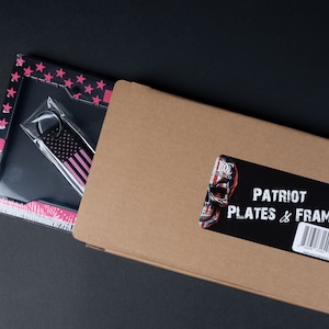 May include: A cardboard box with a sticker that says "PATRIOT PLATES & FRAMES" with a skull graphic. The box is open and a pink and black license plate frame with a matching keychain is visible.
