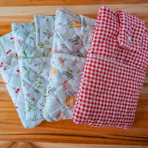 May include: A collection of quilted fabric items, including oven mitts and pot holders, with various floral and gingham patterns. The color palette includes white, red, green, and yellow, with a wooden surface in the background.