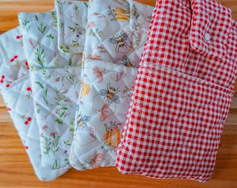 May include: A collection of quilted fabric items, including oven mitts and pot holders, with various floral and gingham patterns. The color palette includes white, red, green, and yellow, with a wooden surface in the background.