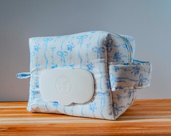 May include: A white and blue floral patterned wipe dispenser bag with a white lid. The bag has a zipper closure and a small side pocket. The bag is sitting on a wooden surface.