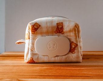 May include: A beige baby wipe dispenser with a zipper closure and a flap. The dispenser is decorated with a repeating pattern of brown teddy bears. The dispenser has a white lid with a butterfly design.