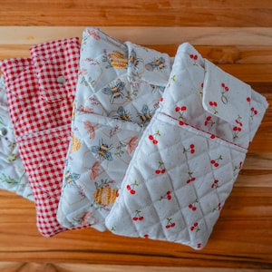 May include: Four quilted fabric pouches with different patterns. One is red and white gingham, another has a floral and bee design, a third has a cherry pattern, and the last has a floral pattern. The pouches are on a wooden surface.