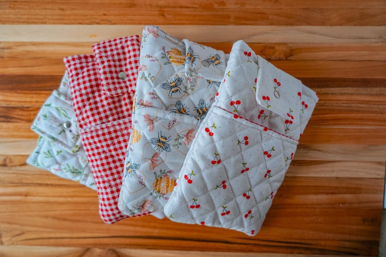 May include: Four quilted fabric pouches with different patterns. One is red and white gingham, another has a floral and bee design, a third has a cherry pattern, and the last has a floral pattern. The pouches are on a wooden surface.