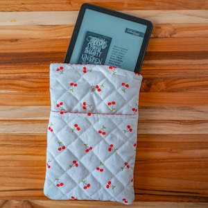 May include: A white quilted tablet sleeve with a cherry pattern. The sleeve has a red trim and a button closure. An e-reader is partially visible inside the sleeve, displaying text from a book.