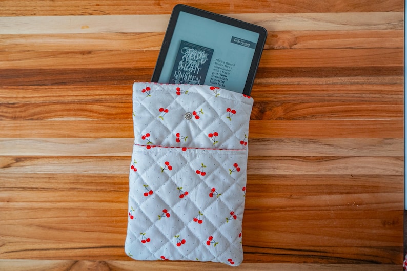May include: A white quilted tablet sleeve with a cherry pattern. The sleeve has a red trim and a button closure. An e-reader is partially visible inside the sleeve, displaying text from a book.