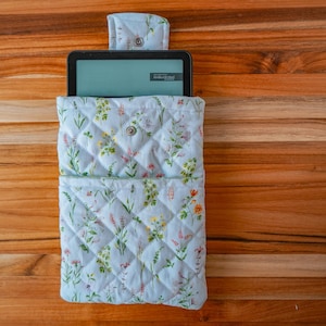 May include: A quilted fabric sleeve for an e-reader featuring a light blue background with a colorful wildflower print. The sleeve includes a front pocket, a flap closure with a metal snap, and a black e-reader device inserted inside.