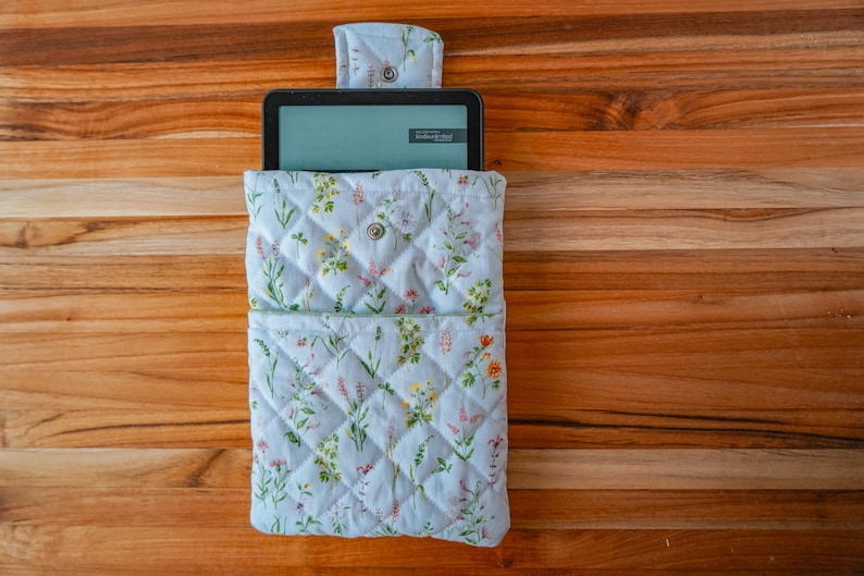 May include: A quilted fabric sleeve for an e-reader featuring a light blue background with a colorful wildflower print. The sleeve includes a front pocket, a flap closure with a metal snap, and a black e-reader device inserted inside.