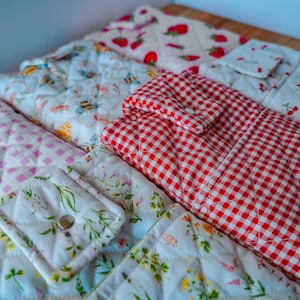 May include: A collection of quilted fabric items with various patterns. Designs include red gingham, strawberries, cherries, bees, and floral prints. The items appear to be kitchen accessories or home decor.