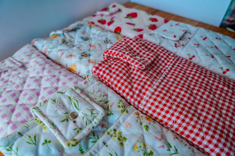 May include: A collection of quilted fabric items with various patterns. Designs include red gingham, strawberries, cherries, bees, and floral prints. The items appear to be kitchen accessories or home decor.