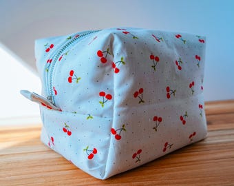 May include: A white, square cosmetic bag with a repeating pattern of red cherries and green leaves. The bag has a silver zipper and a small fabric pull tab. The bag is sitting on a wooden surface.