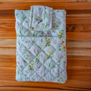 May include: A light blue quilted fabric case with a floral pattern featuring lavender, yellow, and pink flowers. The case has a flap closure with a button and is set on a wooden surface. The case is designed to hold a small item.