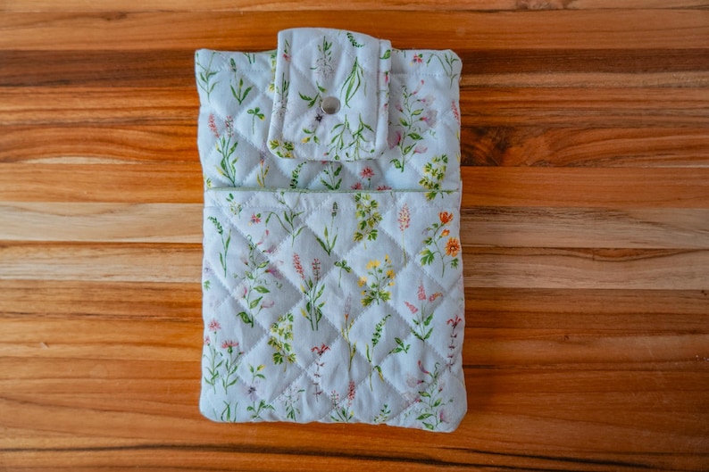 May include: A light blue quilted fabric case with a floral pattern featuring lavender, yellow, and pink flowers. The case has a flap closure with a button and is set on a wooden surface. The case is designed to hold a small item.