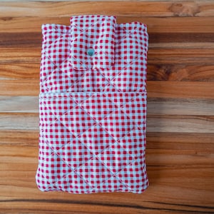 May include: A red and white gingham quilted fabric case with a button closure. The case has a diamond pattern and is designed to hold a tablet or similar device. The case is on a wooden surface.